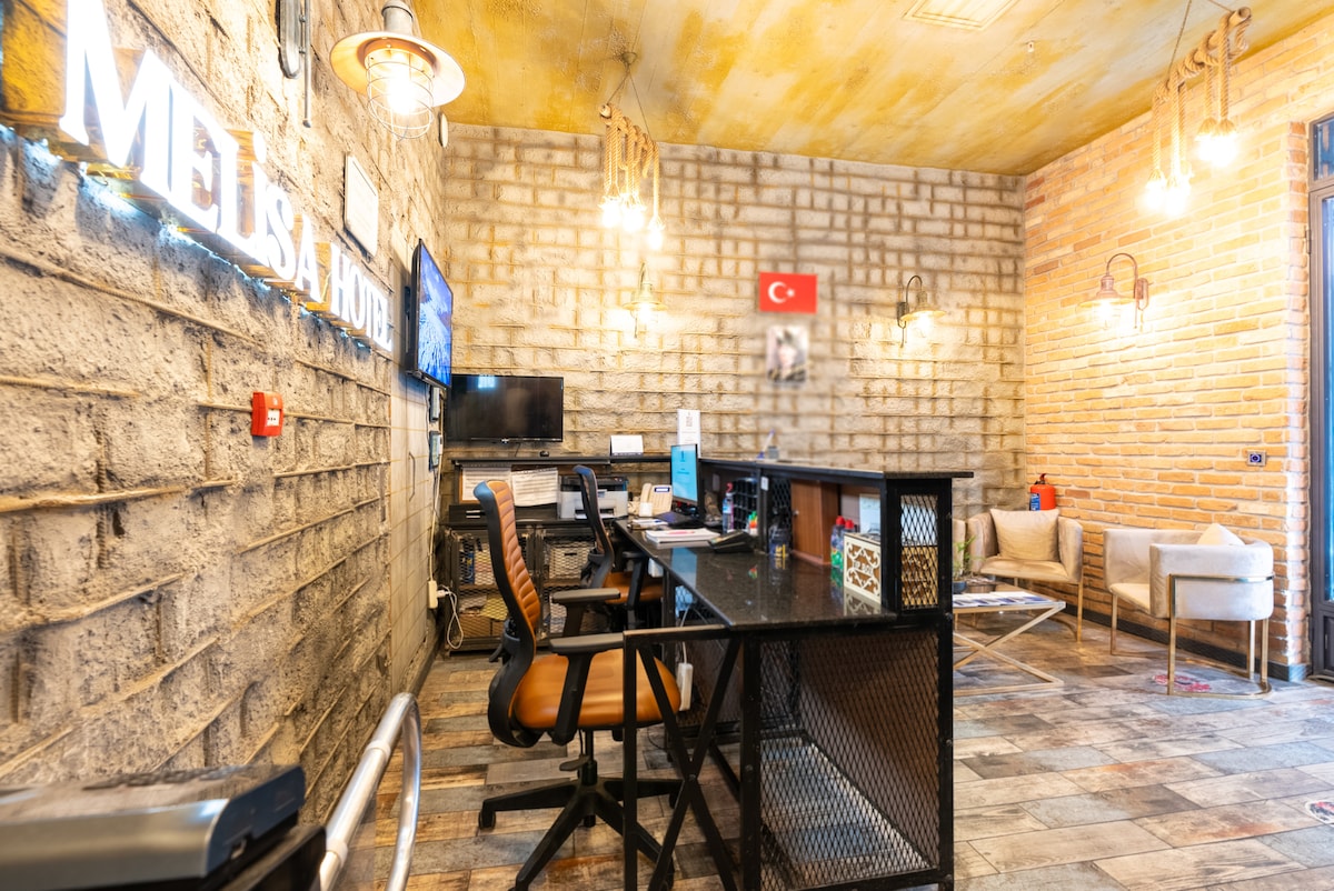 The reception area features a stone wall and wooden ceiling, illuminated by hanging light fixtures. A sleek, dark reception desk equipped with a computer is accompanied by several seating options. A Turkish flag is displayed prominently on the wall, enhancing the welcoming environment.