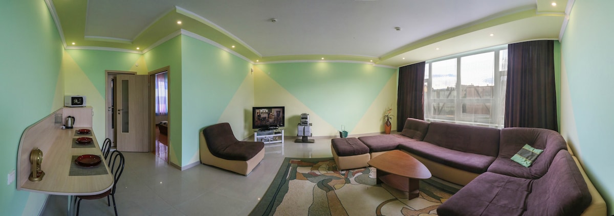 A spacious living area is highlighted by soft green walls and natural light from large windows. A comfortable sectional sofa is positioned around a coffee table, while a television and entertainment unit are visible. A dining setup with chairs is placed in one corner, enhancing functionality.