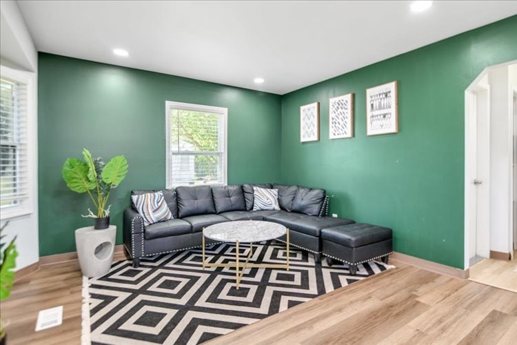 A modern living area features a dark green accent wall, complemented by a large sectional sofa in black leather. A round marble coffee table sits on a patterned black and white rug, and natural light enters through two windows, creating a bright and inviting ambiance.