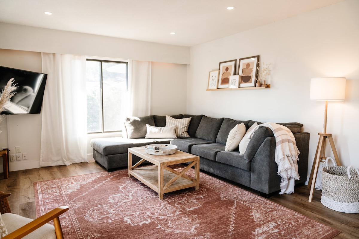 A comfortable living area features a plush sectional sofa adorned with a textured throw and accent pillows. A wooden coffee table sits in front, complemented by a decorative tray. Soft light filters through sheer curtains, illuminating framed art pieces on the wall.
