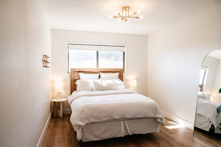 ห้องนอนแขกที่อบอุ่นพร้อมเตียงควีนไซส์ ผ้าปูที่นอนสีขาวสะอาด และหัวเตียงไม้ธรรมชาติ มีไฟข้างเตียงที่ให้แสงนุ่มนวล โคมไฟสีทองทันสมัย และกระจกยาวเต็มตัว การออกแบบที่สว่างและเรียบง่ายเหมาะสำหรับการพักผ่อนยามค่ำคืนอย่างสงบ