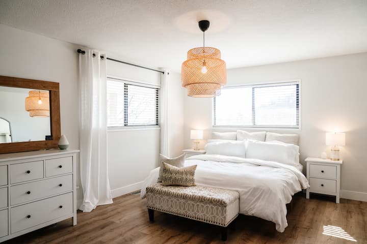 ห้องนอนหลักที่เงียบสงบพร้อมเตียงคิงไซส์หรูหรา ชุดผ้าปูที่นอนสีขาวสะอาด และแสงไฟสีอ่อน มีโคมไฟแขวนแบบสาน ม้านั่งมีสไตล์ ไม้เน้นสีธรรมชาติ และมู่ลี่บังแสง สว่างและโปร่งโล่ง มีหน้าต่างบานใหญ่และการตกแต่งที่เรียบง่ายและสงบ

