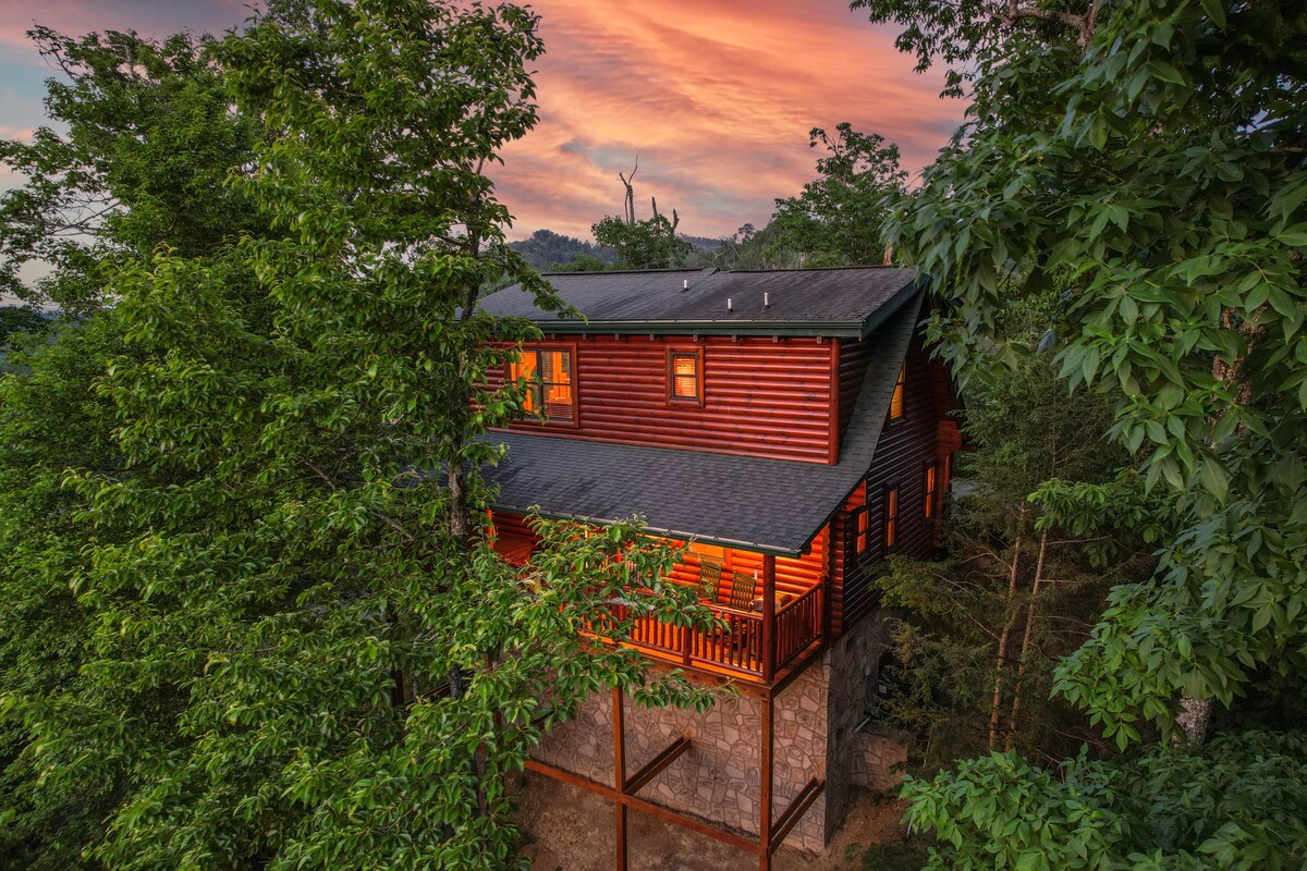 The cabin features a rustic log design, partially obscured by lush greenery. A multi-level structure is highlighted by a wraparound porch, offering scenic views of the surrounding nature and the colorful sky at sunset.