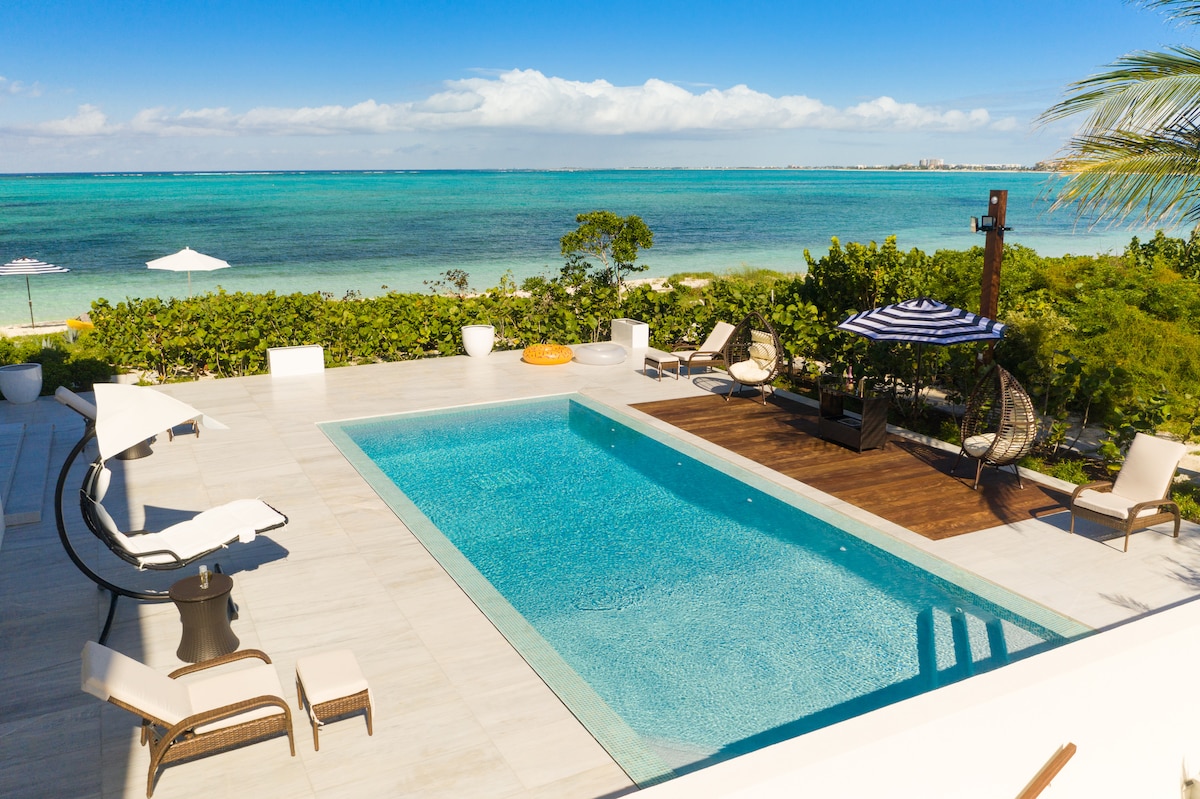 A serene outdoor area features a glimmering swimming pool surrounded by sun loungers and shaded seating. Lush greenery and a view of the turquoise ocean create a tranquil environment, while umbrellas provide shade for relaxation.
