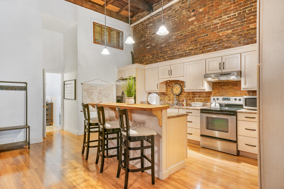 A modern kitchen space features light-colored cabinetry with stainless steel appliances. A wooden breakfast bar with four high-backed stools is positioned in front of the kitchen area. Exposed brick walls add character, while pendant lights illuminate the setting.
