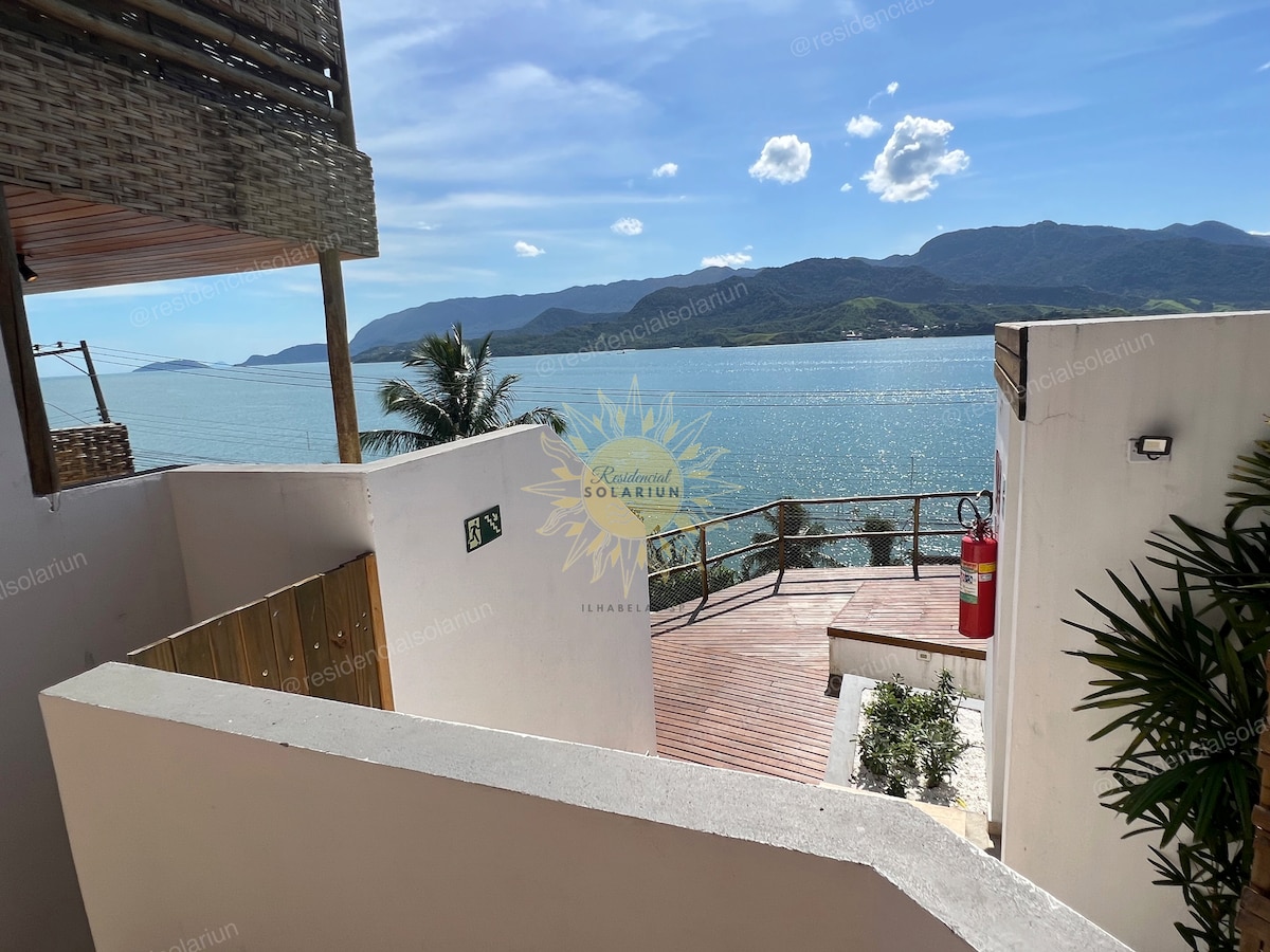 A scenic view of the coastline is presented, featuring tranquil waters reflecting sunlight. Lush greenery and wooden decking create a welcoming outdoor space. The surrounding mountains provide a natural backdrop, enhancing the serene atmosphere of the location.