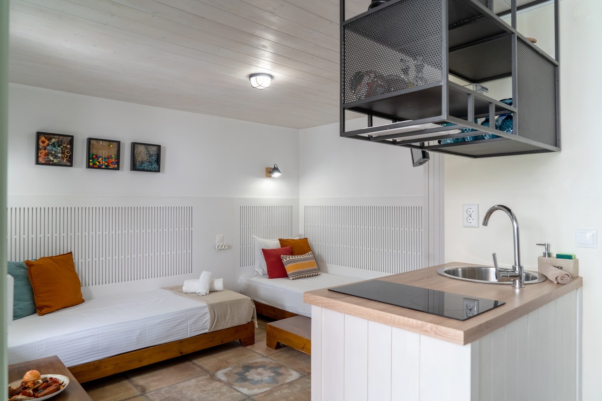 The studio apartment features two single beds accompanied by colorful pillows, situated against white paneling. A well-equipped kitchenette is visible in the corner, complemented by modern shelving. Natural light fills the room, enhancing the functional layout and welcoming atmosphere.