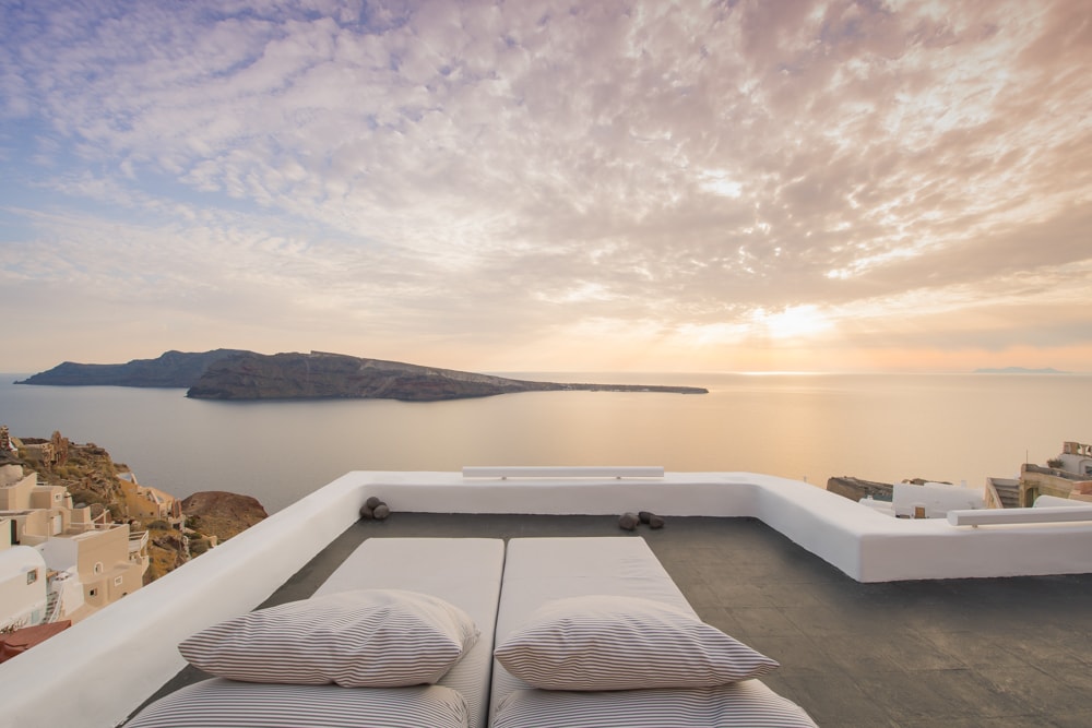A spacious rooftop terrace is featured, equipped with lounge beds positioned for optimal sunset views. The serene landscape includes the Caldera and distant volcanic island, all under a softly colored sky with scattered clouds. The scene invites relaxation in a stunning natural setting.