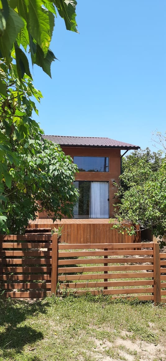 A wooden structure is framed by lush greenery, with a fenced yard showcasing natural landscaping. Large windows invite light into the upper-level space, while a balcony extends outward, providing a serene outdoor area. The clear blue sky enhances the inviting exterior.