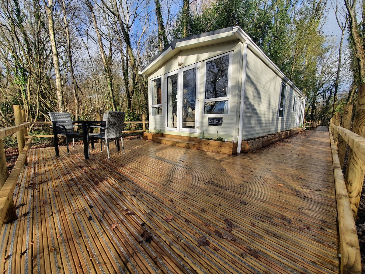 A spacious deck area frames the lodge, constructed from wooden planks and surrounded by greenery. Two outdoor chairs and a small table invite relaxation, while large glass doors offer a view into the interior. Trees provide a natural backdrop, enhancing the lodge's secluded setting.