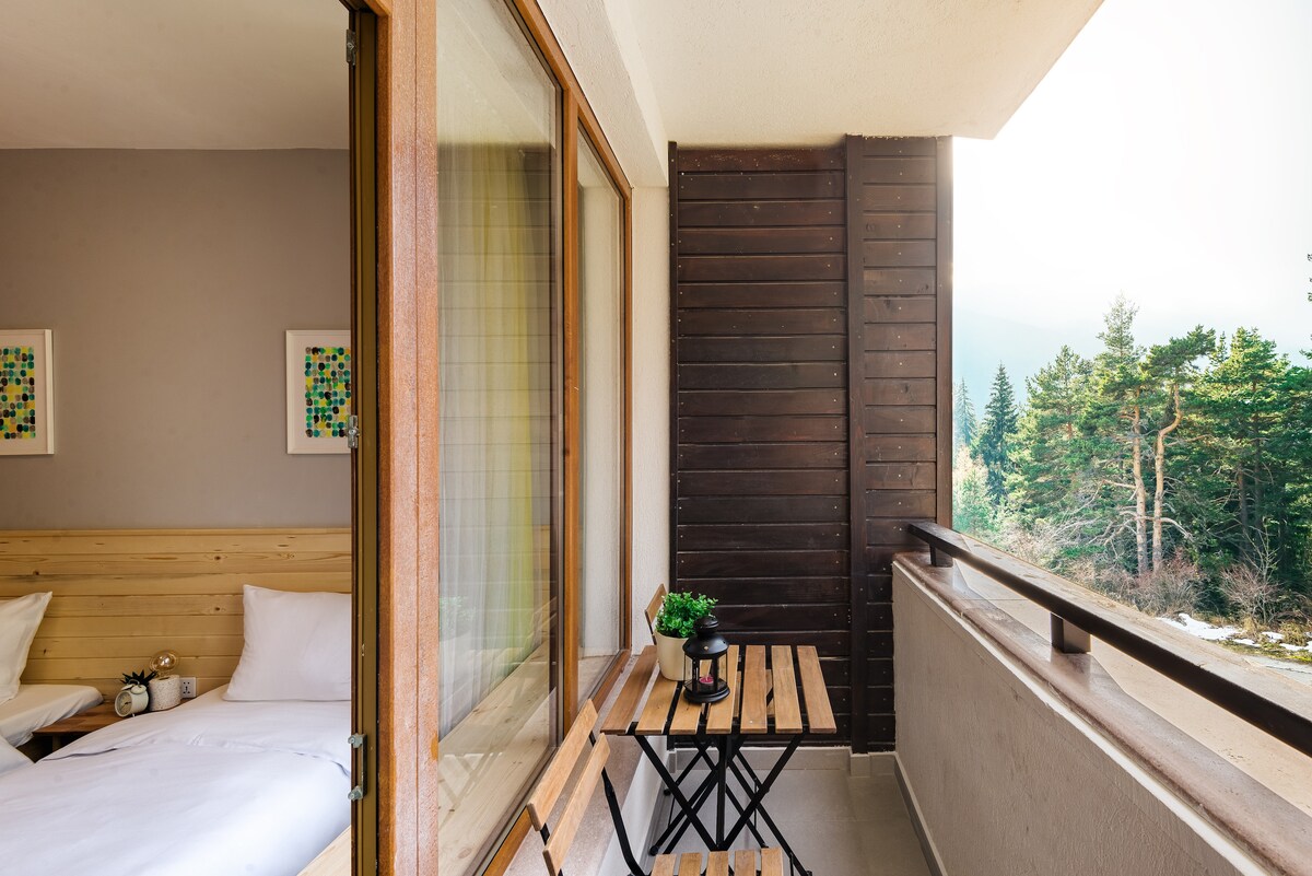 A private balcony is viewed with a wooden table and a contrasting black folding chair. Light-colored walls and natural wood accents create a warm setting. Beyond, trees and mountains are visible, enhancing the tranquil atmosphere of the space.