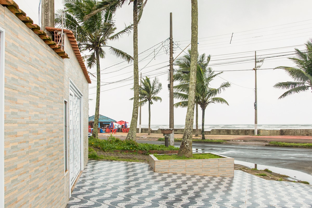 A tiled area is positioned outside the house, featuring distinct patterns. Towering palm trees are visible along the street, with the beach seen in the distance. A few colorful kiosks can be noticed across the road, contributing to the beachside setting.