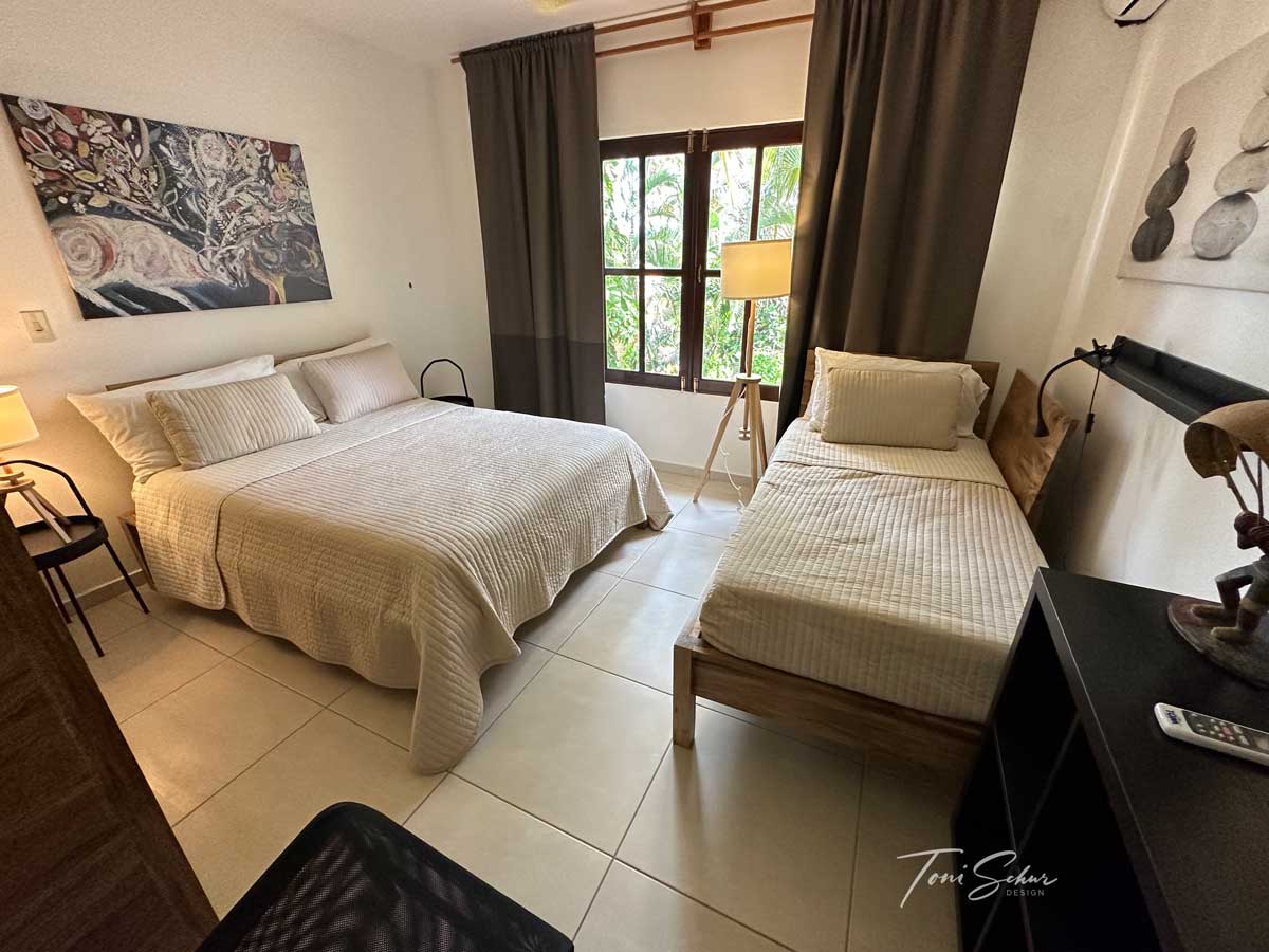 The bedroom features a spacious bed and a single bed, accompanied by a bedside table and a lamp. Natural light filters through the window adorned with dark curtains, providing a serene atmosphere. Artwork is displayed on the wall, enhancing the inviting space.