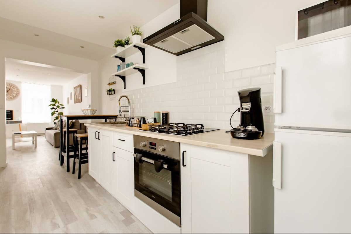 The kitchen features modern appliances including a gas stove and an oven, complemented by sleek white cabinetry. A coffee maker sits alongside a refrigerator, with ample counter space and decorative shelf plants enhancing the functional design. The dining area is visible in the background.