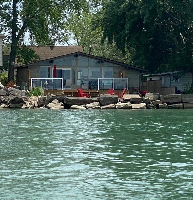 A waterfront cottage is situated on rocky terrain, featuring a large deck with red Muskoka chairs. Large windows allow sunlight to fill the interior. Surrounding greenery creates a natural environment, enhancing the tranquil setting.