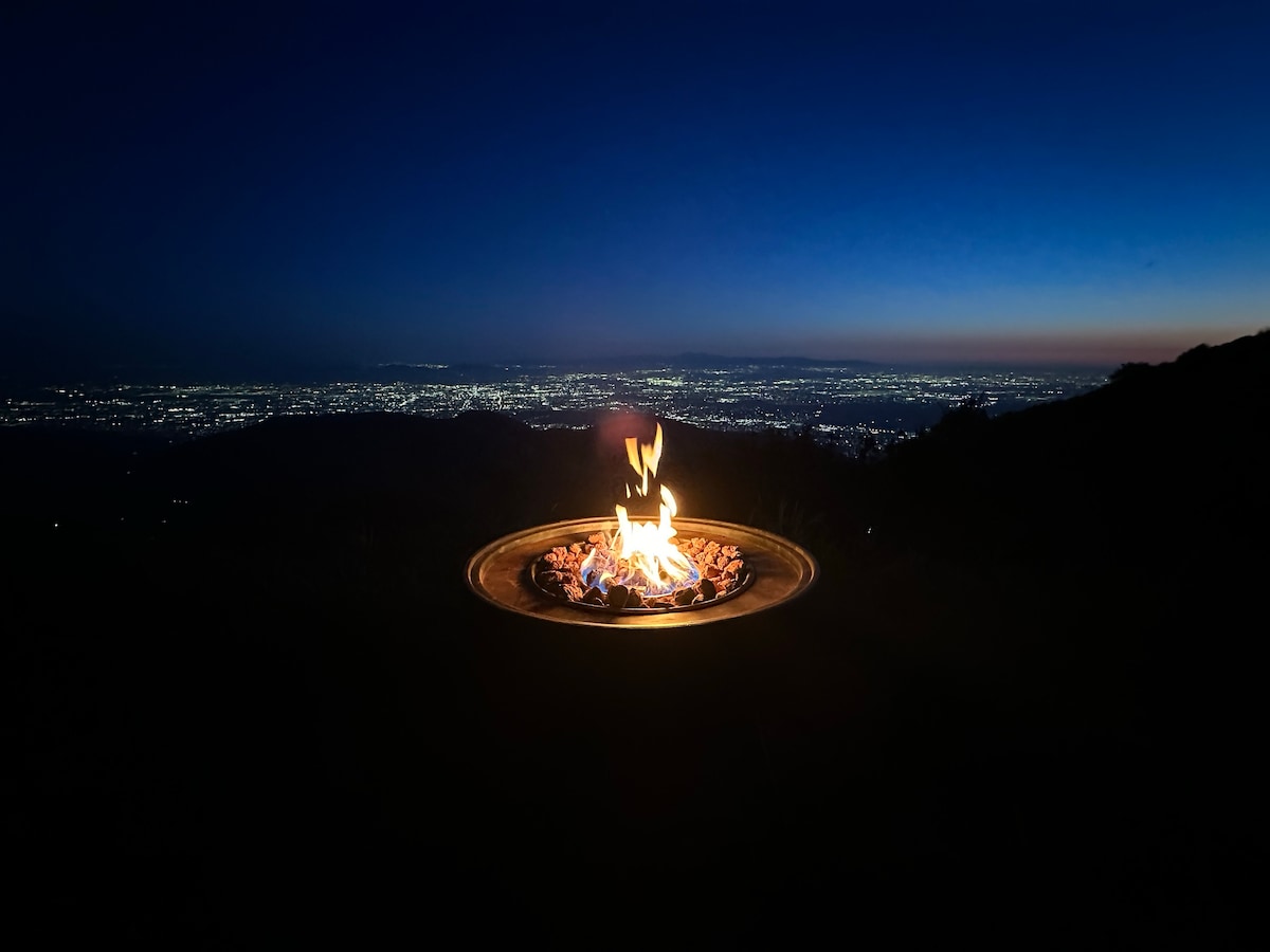 A fire pit, prominently featured in the foreground, glows warmly as flames dance above glowing embers. The backdrop reveals a panoramic view of the city, adorned with twinkling lights transitioning from dusk to evening, creating a serene ambiance.