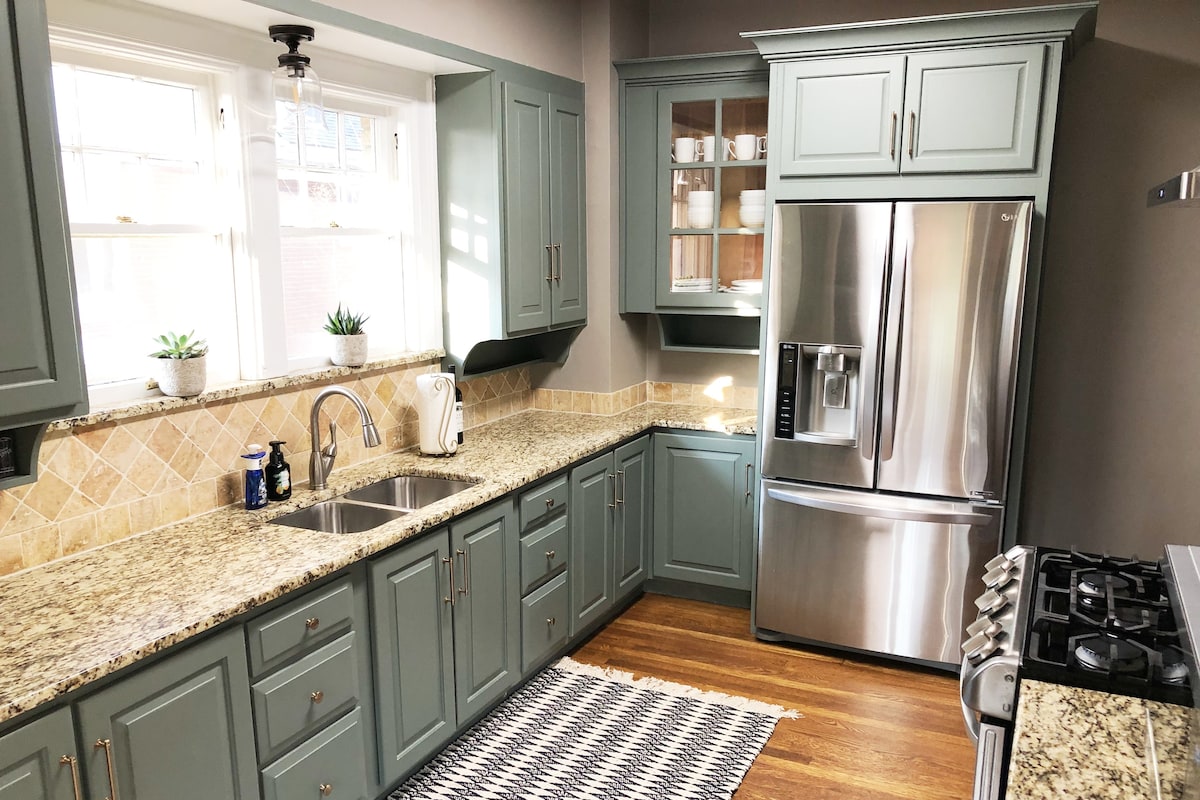 The kitchen features modern cabinetry in a soft green hue, complemented by granite countertops and a stylish tile backsplash. Stainless steel appliances are present, including a refrigerator and a gas oven. Natural light filters through two windows adorned with plants, enhancing the inviting atmosphere.