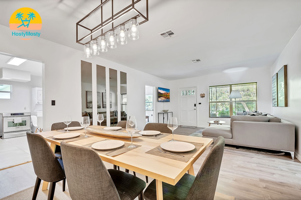 A modern dining area features a spacious wooden table set for six, complete with dinnerware and glassware. Soft natural light brightens the room, highlighting the contemporary lighting fixture above. The adjacent living area is visible, showcasing a comfortable sofa and wall-mounted television.
