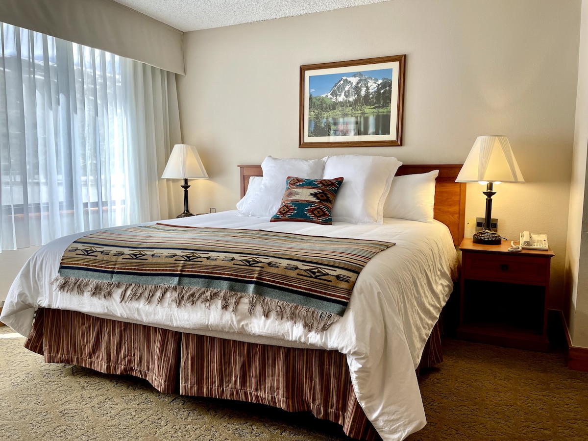 A spacious king bed is dressed in crisp white linens, complemented by a patterned throw. Two nightstands with lamps are positioned on either side. A framed mountain landscape hangs above the bed, with natural light filtering through sheer curtains.