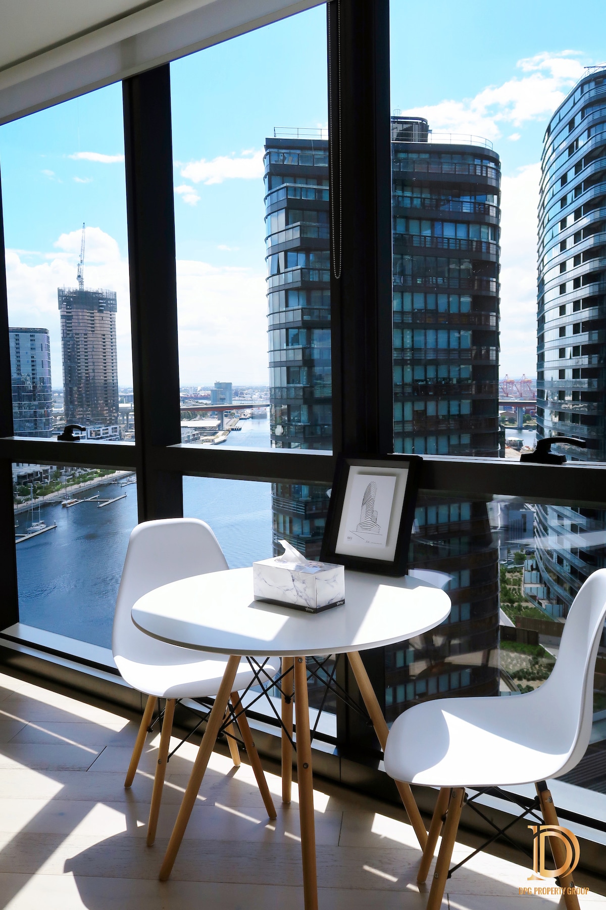 A small dining area is positioned by a large window, offering a view of the surrounding buildings and water. A round table is paired with two sleek white chairs. A framed artwork and a tissue box are displayed on the table, enhancing the space's functionality.
