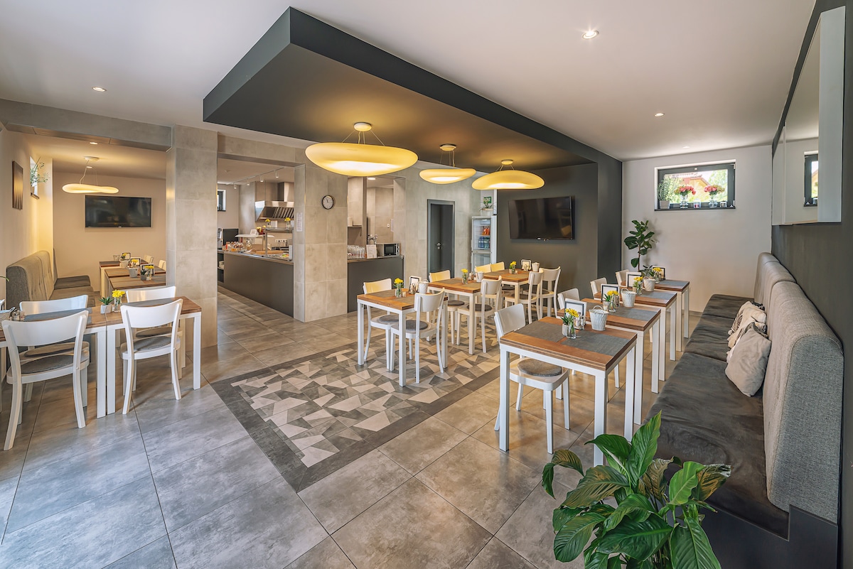 The dining area features a modern design with light wooden tables and white chairs arranged in rows. A gray and white patterned rug anchors the space, while a large kitchen is visible in the background. Soft overhead lighting adds to the ambiance.