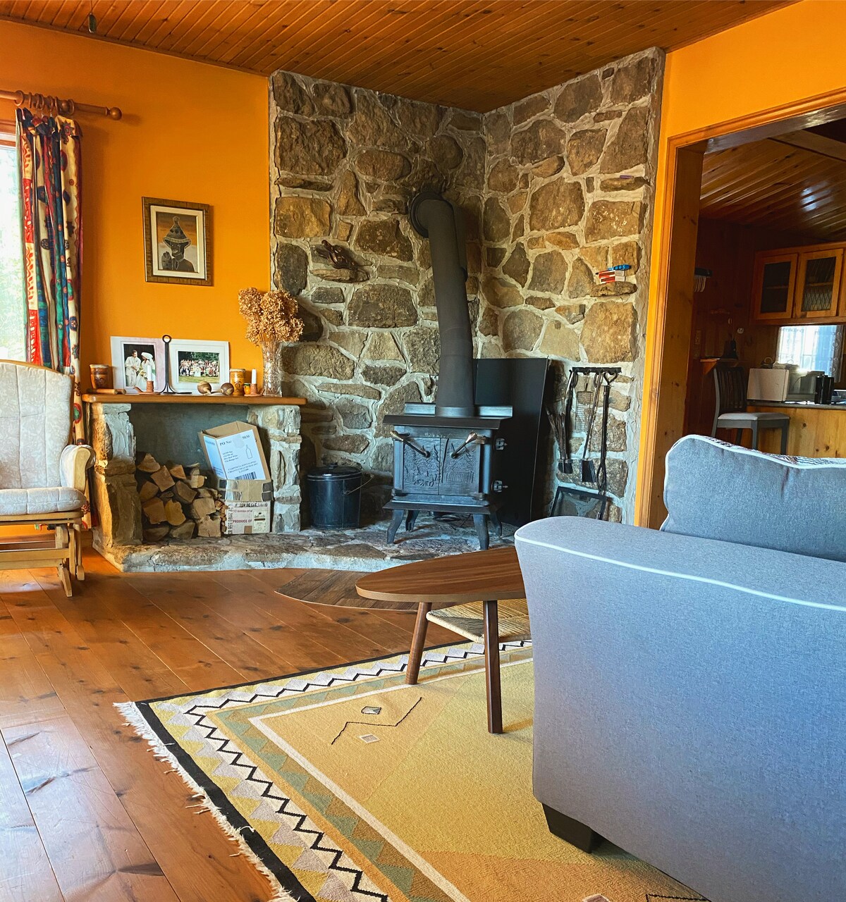 A cozy living area features a stone fireplace, complemented by a wood stove and neatly stacked firewood. Colorful curtains frame a window, while a small coffee table sits on an area rug. Warm wood tones create a welcoming ambiance throughout the space.