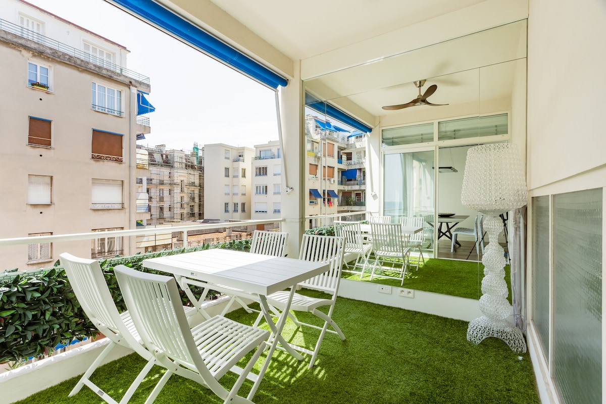 A spacious terrace is furnished with a white dining table and several white chairs, surrounded by lush greenery. Large glass doors provide natural light and an open view of the neighboring buildings, while a decorative lamp and ceiling fan add modern touches.