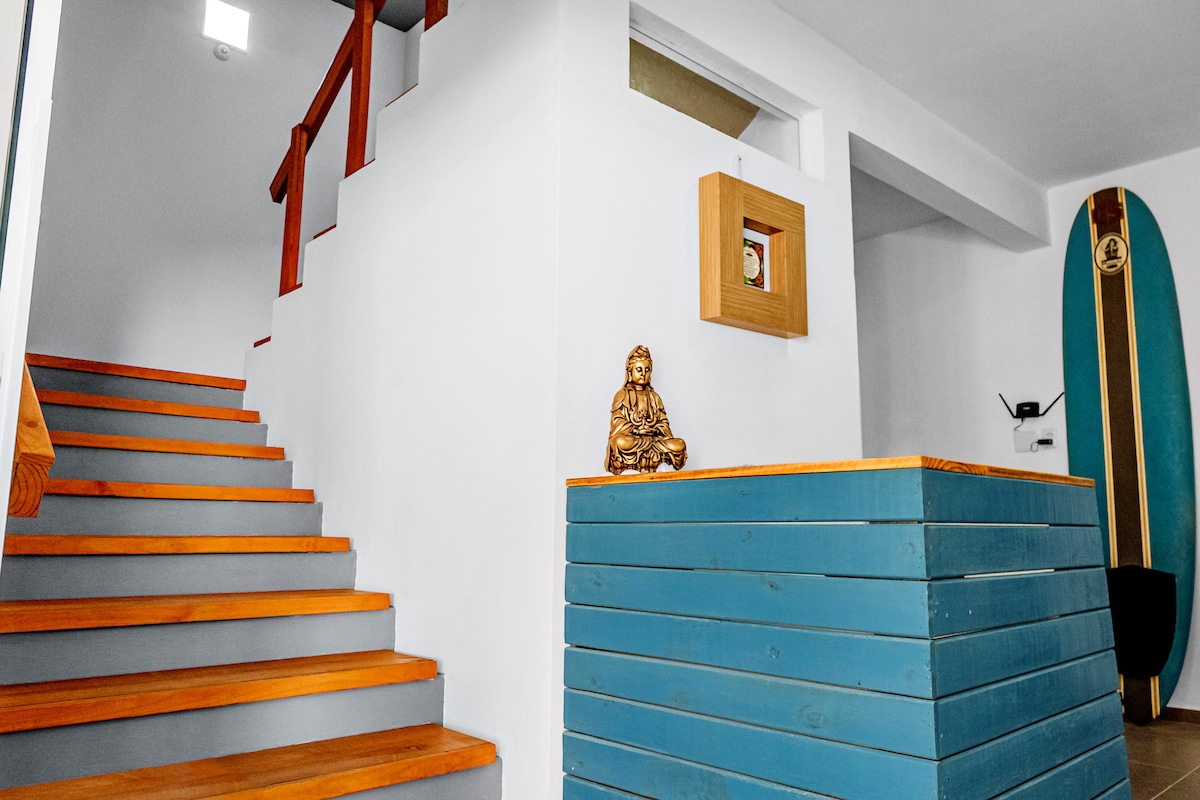 An interior view showcases a staircase with wooden steps leading to an upper floor. A blue partition is visible at the base of the stairs, with a decorative frame and a small statue. A surfboard rests against the wall, contributing to a relaxed ambiance.