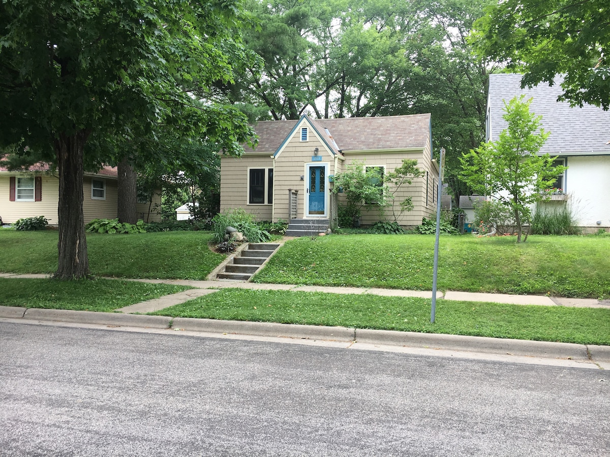 A single-story home is set back from the street, featuring a light beige exterior and a blue front door. The yard is adorned with greenery and small plants, while steps lead up to the entrance. Mature trees provide shade in the surrounding area.