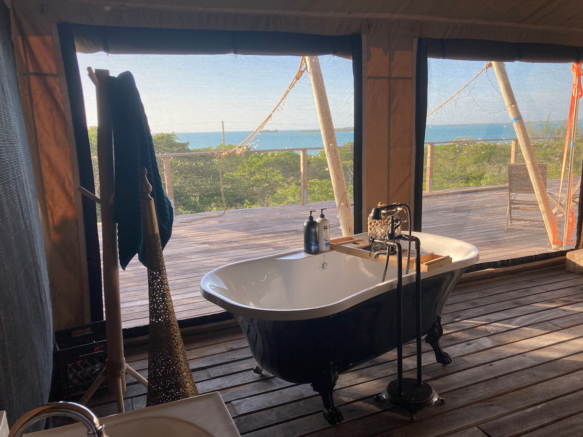 A freestanding black and white bathtub is positioned in front of large, open canvas walls, providing views of the lush landscape and coastline beyond. The wooden deck features a natural finish, complementing the earthy tones of the space.