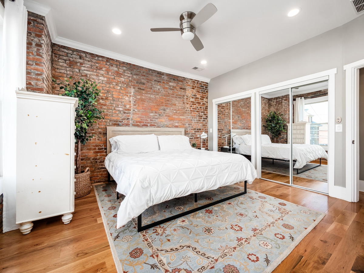 Charmigt sovrum med en exponerad tegelvägg, trägolv och en bekväm queen-size säng. Utrymmet är inrett med en mönstrad matta, en takfläkt och ett vitt förvaringsskåp, och det blir ljusare av en touch av grönska som ger en mysig, inbjudande känsla.