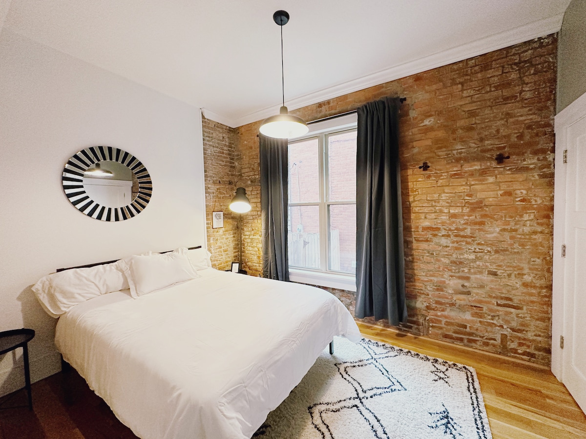 Mysigt sovrum med en exponerad tegelvägg, bekväma sängkläder och en elegant spegel – skapar ett varmt och inbjudande utrymme, perfekt för en avkopplande vistelse.