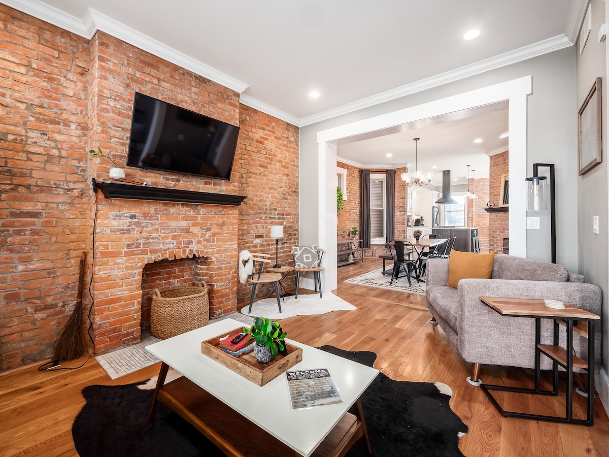 Charmigt och rymligt vardagsrum med exponerade tegelväggar, en mysig öppen spis och bekväma sittplatser – perfekt för avkoppling. Njut av elegant inredning, en stor TV och gott om naturligt ljus i hela utrymmet.