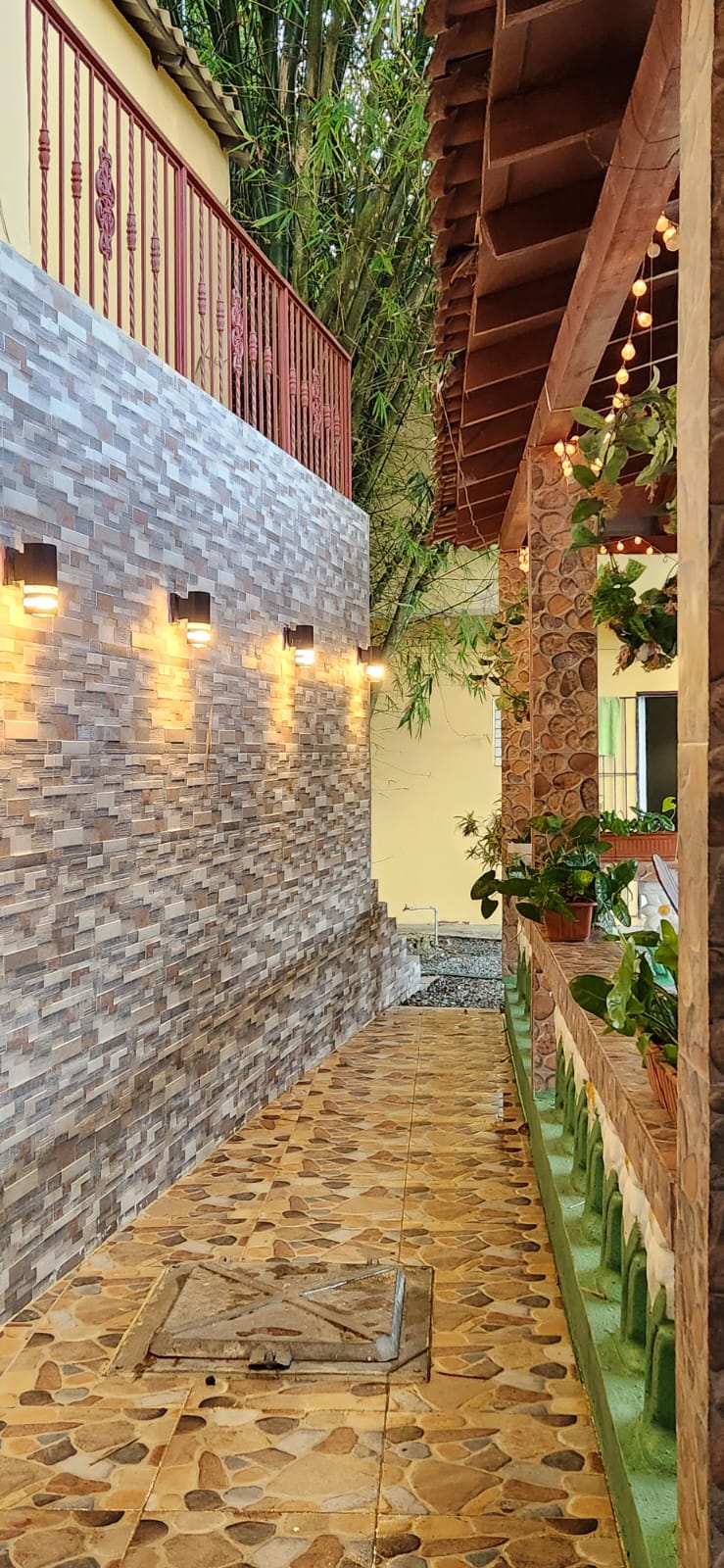 A narrow walkway is flanked by textured stone walls and is softly lit by wall-mounted lights. Natural greenery is integrated throughout the space, providing a fresh ambiance. Decorative tiles in earthy tones cover the ground, leading to an inviting entrance area adorned with potted plants.