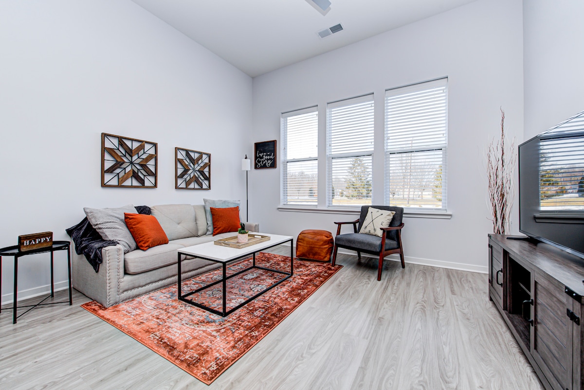 A modern living area features a light-colored couch adorned with orange throw pillows. A coffee table is positioned in front, complemented by a decorative area rug. Large windows allow natural light to fill the space, with a cozy armchair added for seating.