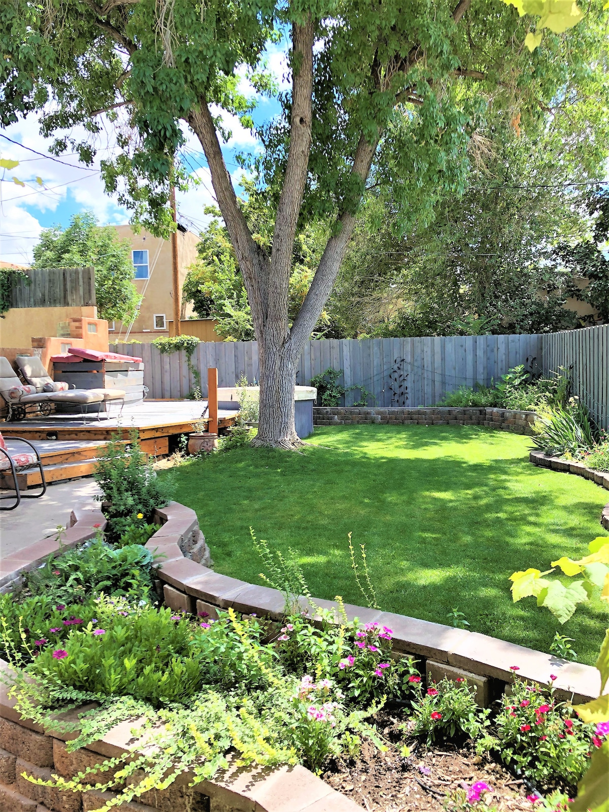 A well-maintained backyard features a lush green lawn surrounded by vibrant flower beds. A patio area with comfortable seating and a BBQ grill is positioned under the shade of a large tree, creating a serene outdoor space. A wooden fence offers privacy.