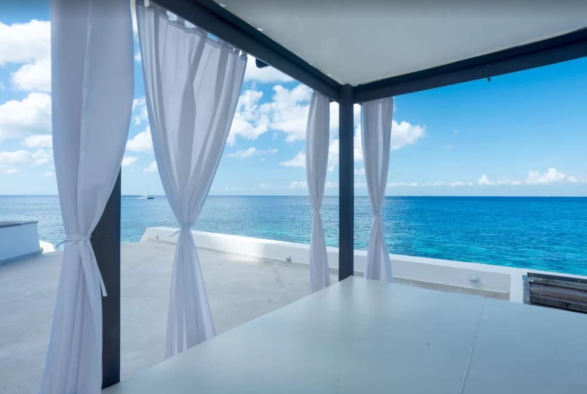 The image showcases an oceanfront view from a shaded outdoor seating area. Flowing white curtains frame the space, accentuating the bright blue sky and deep blue Caribbean waters. A smooth, light-colored surface extends beyond, providing a tranquil ambiance.