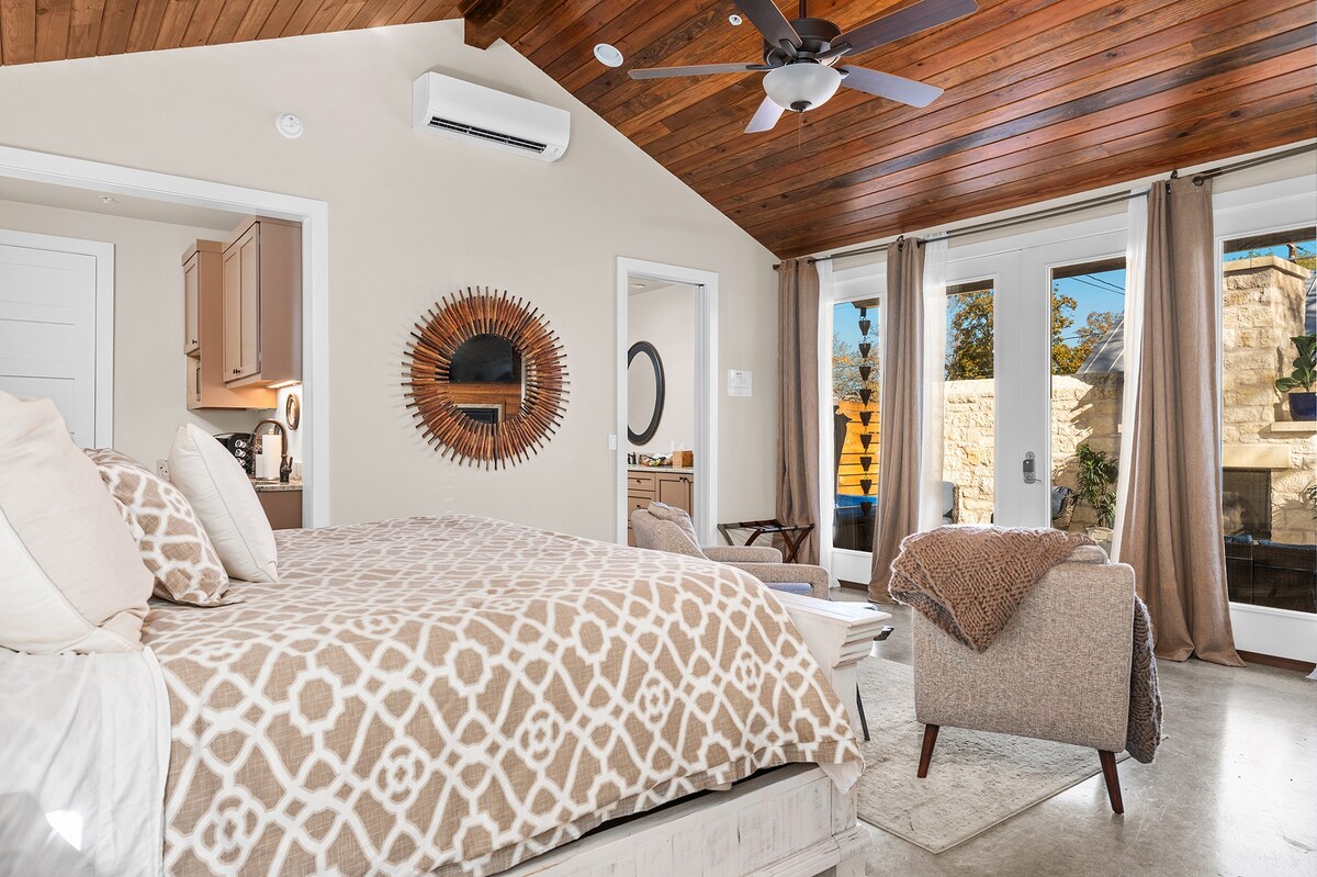 A spacious bedroom features a king bed with a patterned blanket and decorative pillows. A ceiling fan and a large mirror enhance the aesthetic. Natural light enters through glass doors leading to an outdoor area, revealing a peaceful retreat atmosphere.