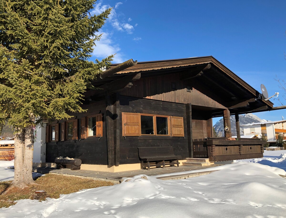 The chalet features a traditional wooden exterior with deep brown tones and decorative shutters. A spacious front porch is visible, accompanied by seating and a surrounding snow-covered yard. A tall conifer tree adds to the serene, wintry landscape under a clear blue sky.