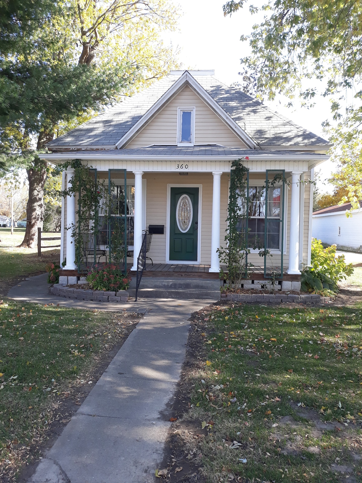 A charming home is presented with a light-colored facade and a green front door. Climbing vines adorn the porch columns, and the landscaped front yard features a path leading to the entry. Tall trees provide shade around the property.
