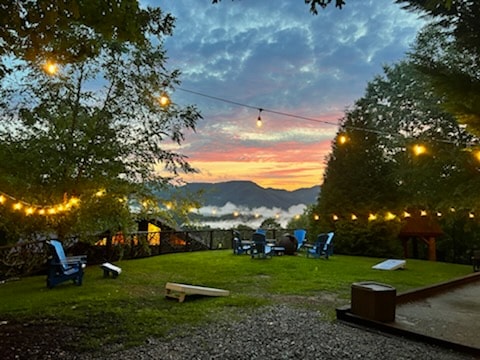 An outdoor area is illuminated by string lights, enhancing the evening ambiance. Several blue chairs are positioned on a grassy patch, overlooking a scenic view of mountains and a cloudy valley during sunset.