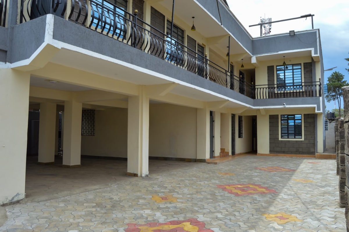 An expansive outdoor area is shown, with a patterned stone pavement featuring colorful designs. The ground floor of the building is visible, showcasing a balcony with decorative railings above. Large windows provide natural light to the interior spaces.