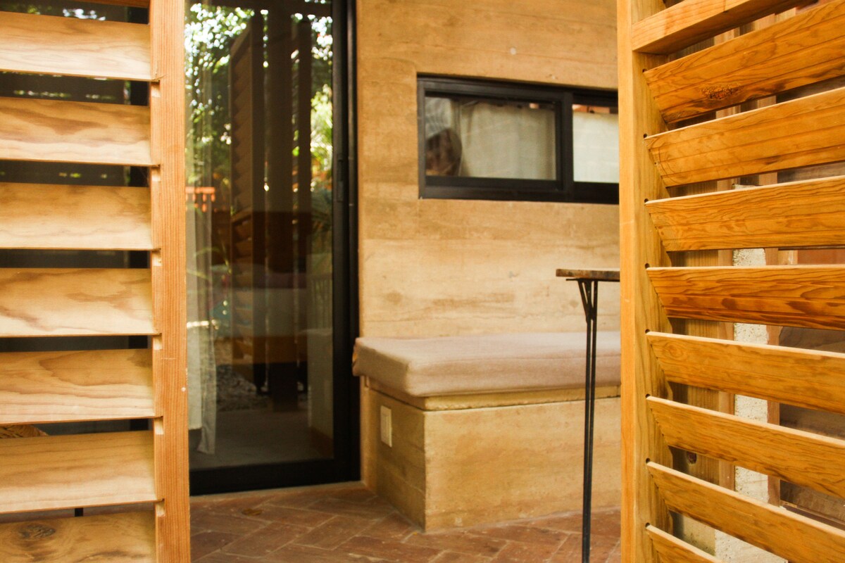 A shaded seating area is visible under natural wood slats, leading to a tranquil space with a low bench and a small table. Large glass doors reflect the surrounding greenery, providing a connection to the outdoors.