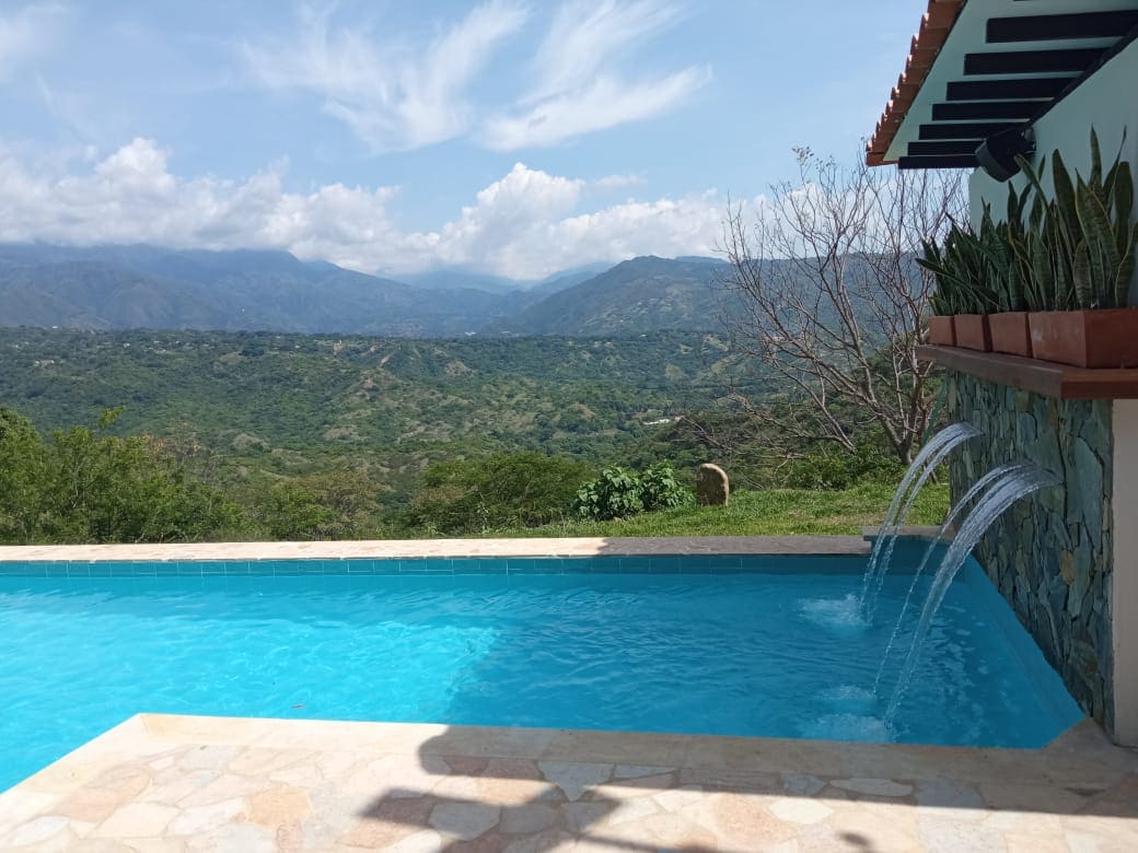 A refreshing pool is featured with three water fountains, providing a serene ambiance against a backdrop of rolling green hills and distant mountains. The clear water reflects the sky, enhancing the outdoor space for relaxation and enjoyment.