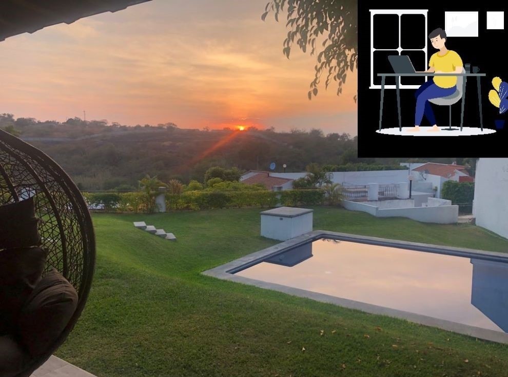 A serene outdoor view features a well-maintained lawn leading to a swimming pool that reflects the sunset. The backdrop includes gentle hills and neighboring homes. A comfortable seating area is visible, providing an inviting space for relaxation or work.