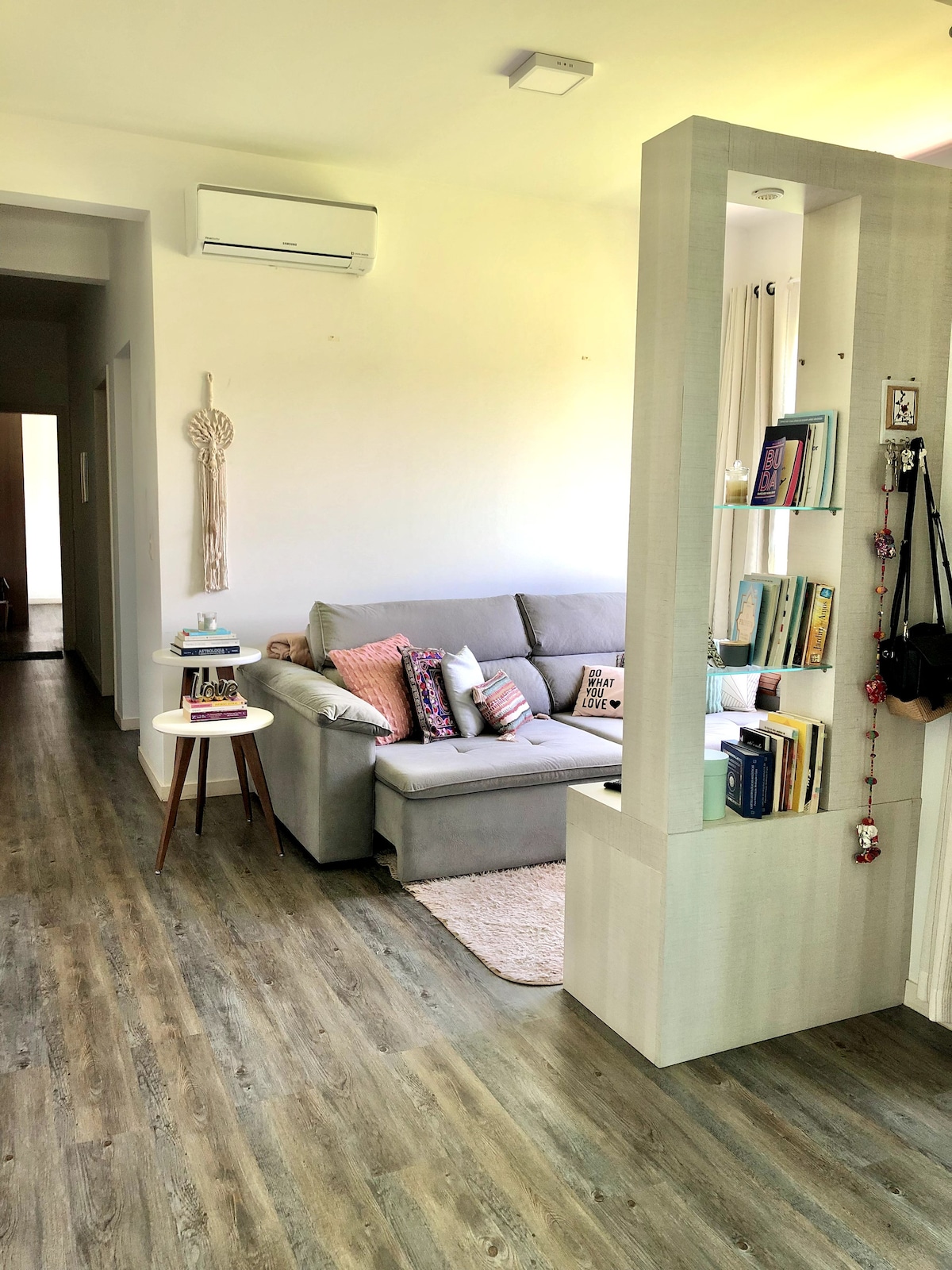A modern living area is depicted with a comfortable gray sofa adorned with colorful cushions. A small white side table sits next to the sofa, while a bookshelf visually separates the space, showcasing various books and decorative items. Natural light illuminates the room from the front.