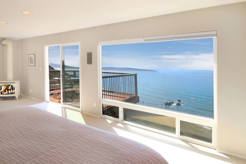 A spacious bedroom features a large floor-to-ceiling window, showcasing sweeping ocean views. Natural light fills the room, and a deck is accessible through sliding glass doors. A subtle fireplace adds a cozy touch to the serene atmosphere.