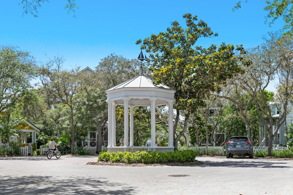 A white gazebo with a pointed roof is positioned centrally in the image, surrounded by lush greenery. A bicycle and a parked car are visible on a paved path leading through the area, demonstrating a welcoming community space.