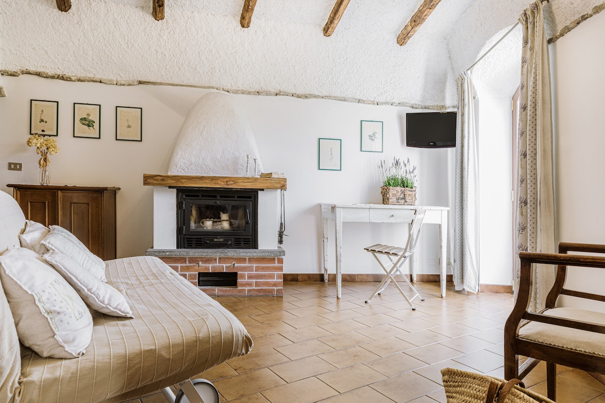 A cozy living area is highlighted by a traditional fireplace with a wooden mantle. A sofa bed and a small table are present, complemented by a few framed artworks on the walls. Natural light enters through a partially open door, illuminating the terracotta tiled floor.