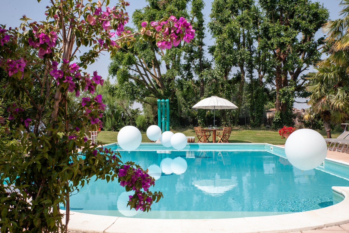 A tranquil swimming pool is surrounded by lush greenery, with vibrant flowers adding color to the scene. Inviting white spheres float on the water’s surface, and sun loungers are arranged under a large umbrella, creating a relaxing outdoor space.